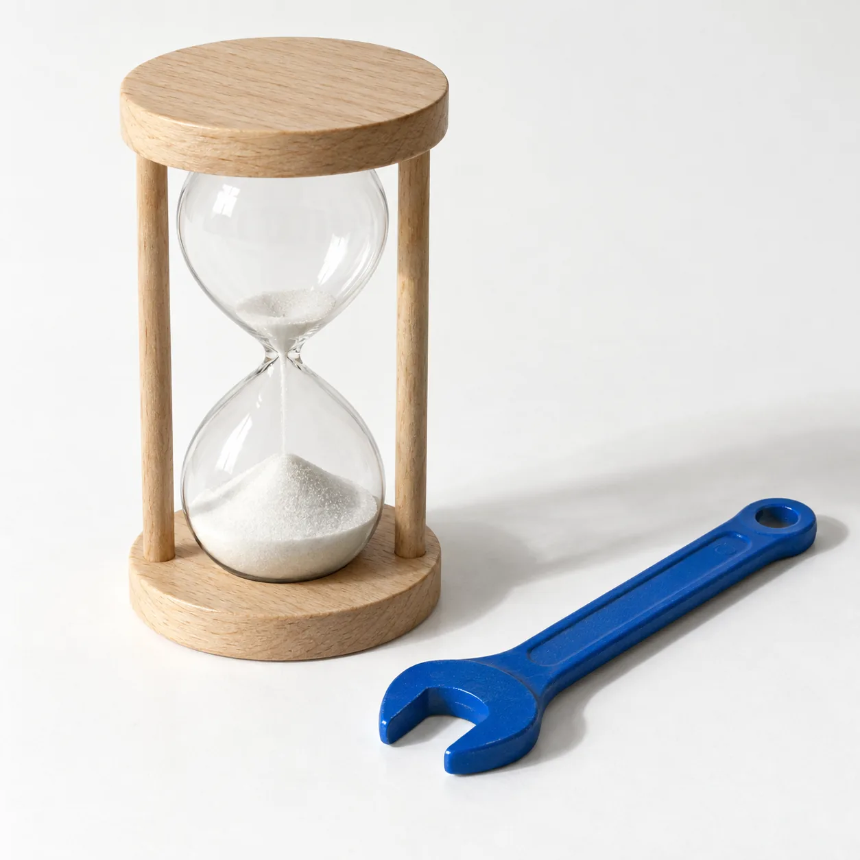 Eine Sanduhr mit weißem Sand, deren oberer Kolben fast leer ist, daneben liegt ein blauer Schraubenschlüssel bereit.